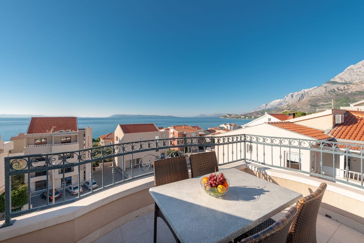 Ein Balkon mit Tisch und Stühlen, einer Obstschale und Blick auf das Meer und Gebäude.