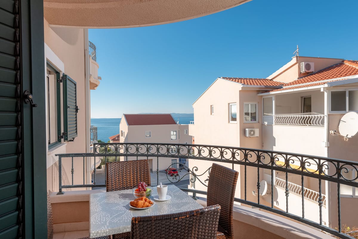 Ein Balkon mit Korbstühlen, einem Tisch mit Obstschale und Tasse, und Blick auf das Meer und Gebäude.