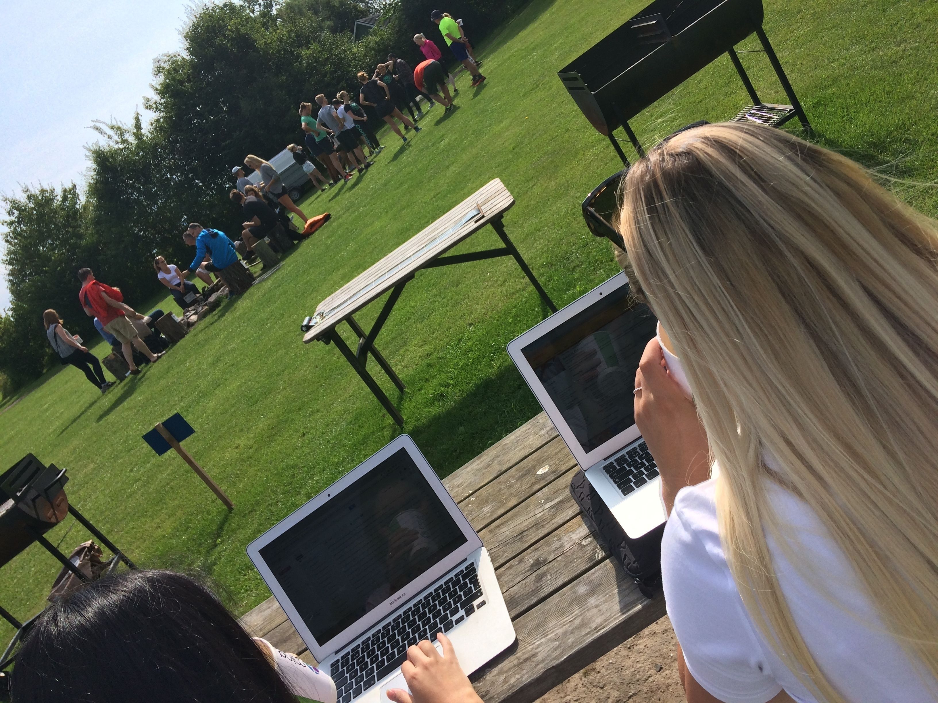 Een groep mensen buiten, laptops op een houten tafel, wat wijst op een informele vergadering of bijeenkomst.