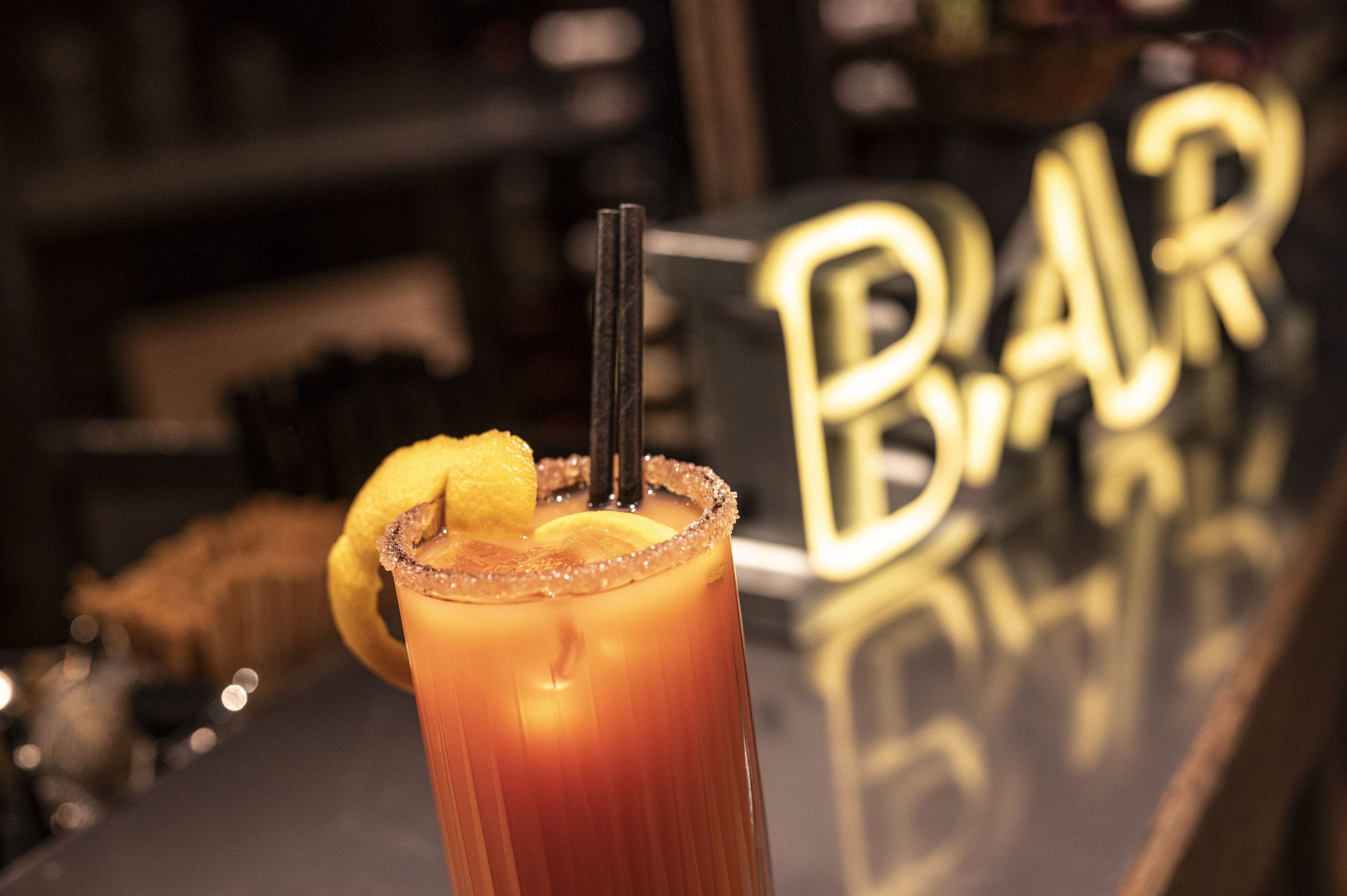 A close-up of a Bloody Mary cocktail with a lemon slice garnish and a salted rim, placed on a bar counter with a blurred background featuring a sign that reads "BAR".
