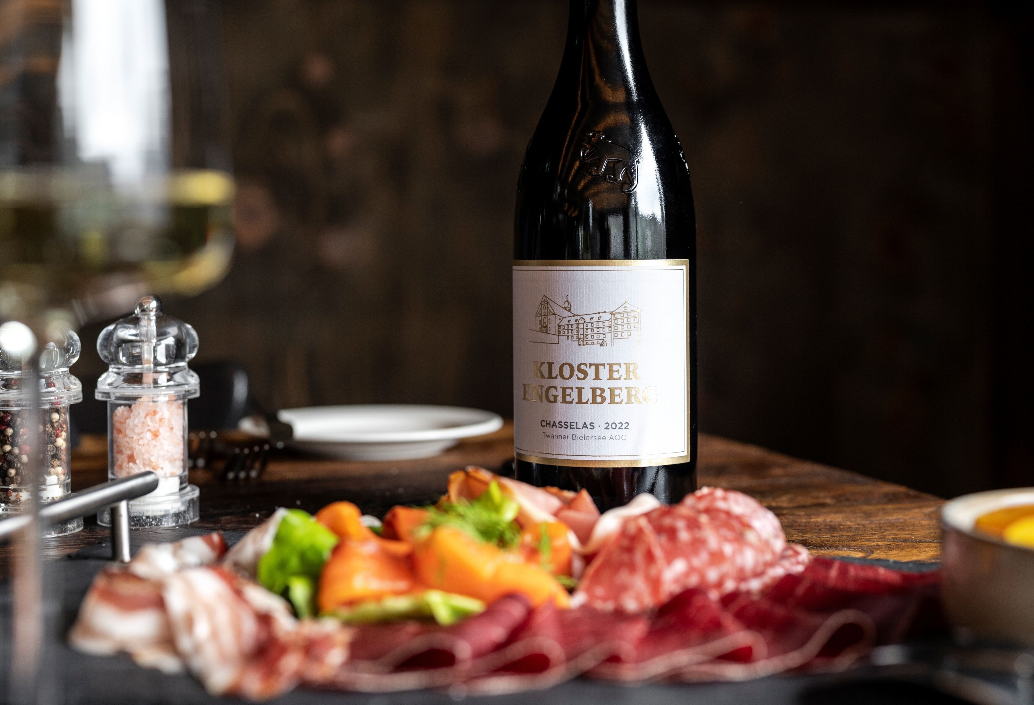 Una botella de vino Kloster Engelberg, una mesa de madera con diversas carnes y verduras, además de saleros y pimenteros.