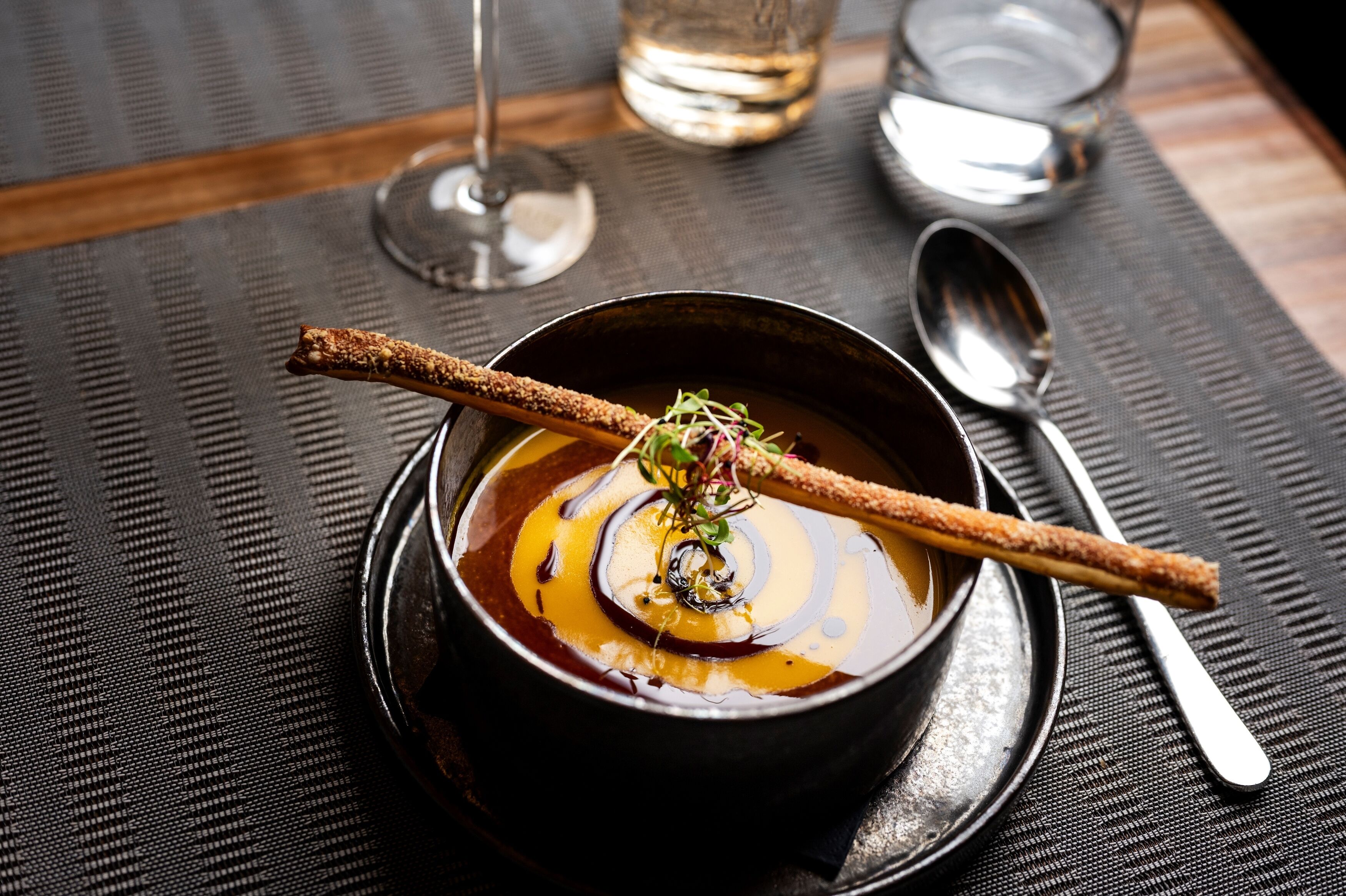A bowl of soup with a decorative stick and garnish on a dark plate, accompanied by a spoon and a glass of water on a textured tablecloth.