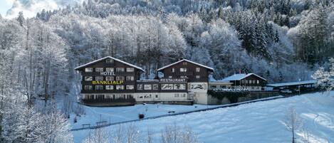 Un hotel con restaurante y un edificio bancario, rodeado de árboles y montañas cubiertas de nieve.