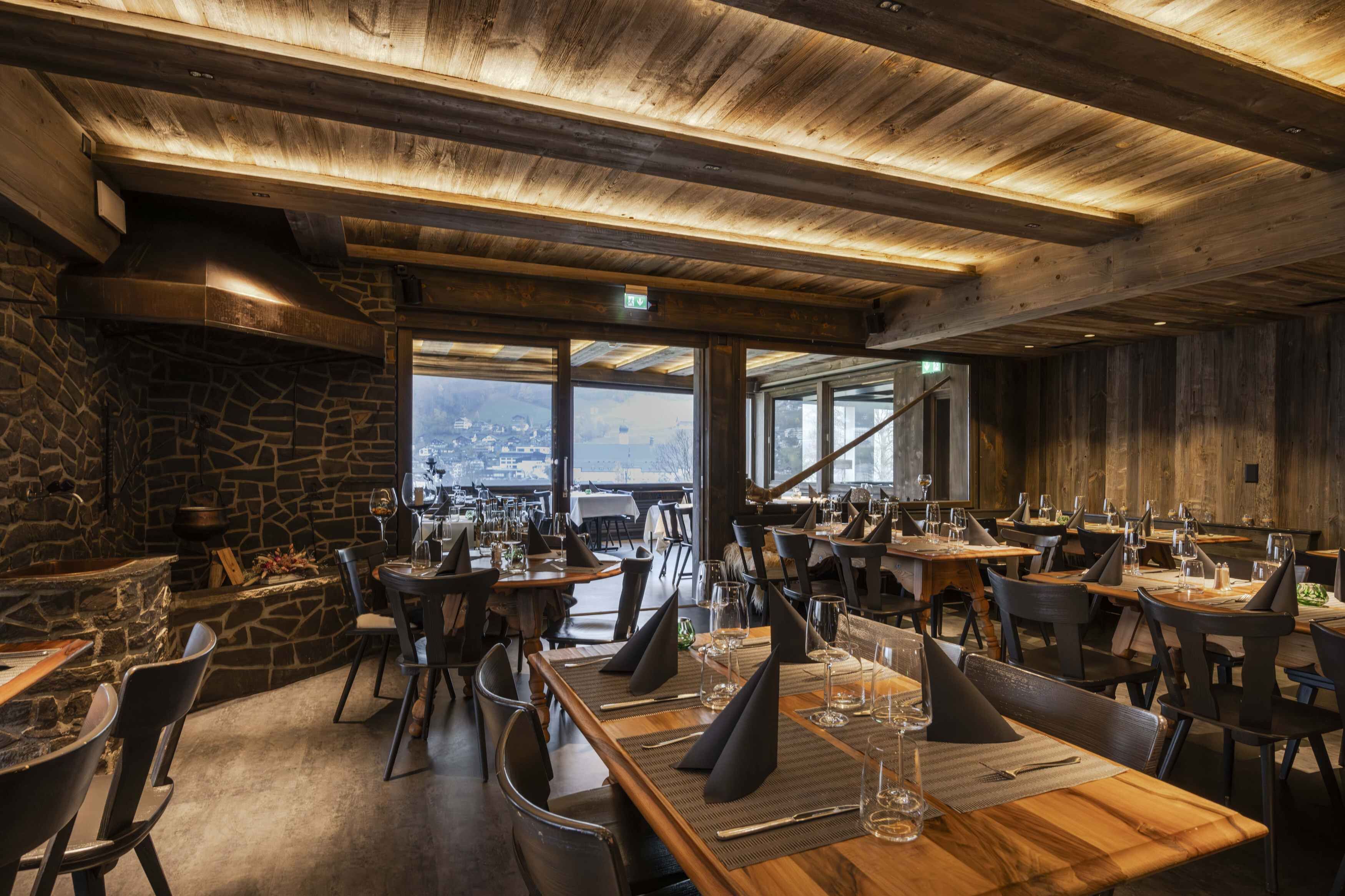 Un restaurante con un interior rústico, paredes de piedra, vigas de madera y amplios ventanales que ofrecen vistas. Las mesas están preparadas con copas de vino y servilletas.