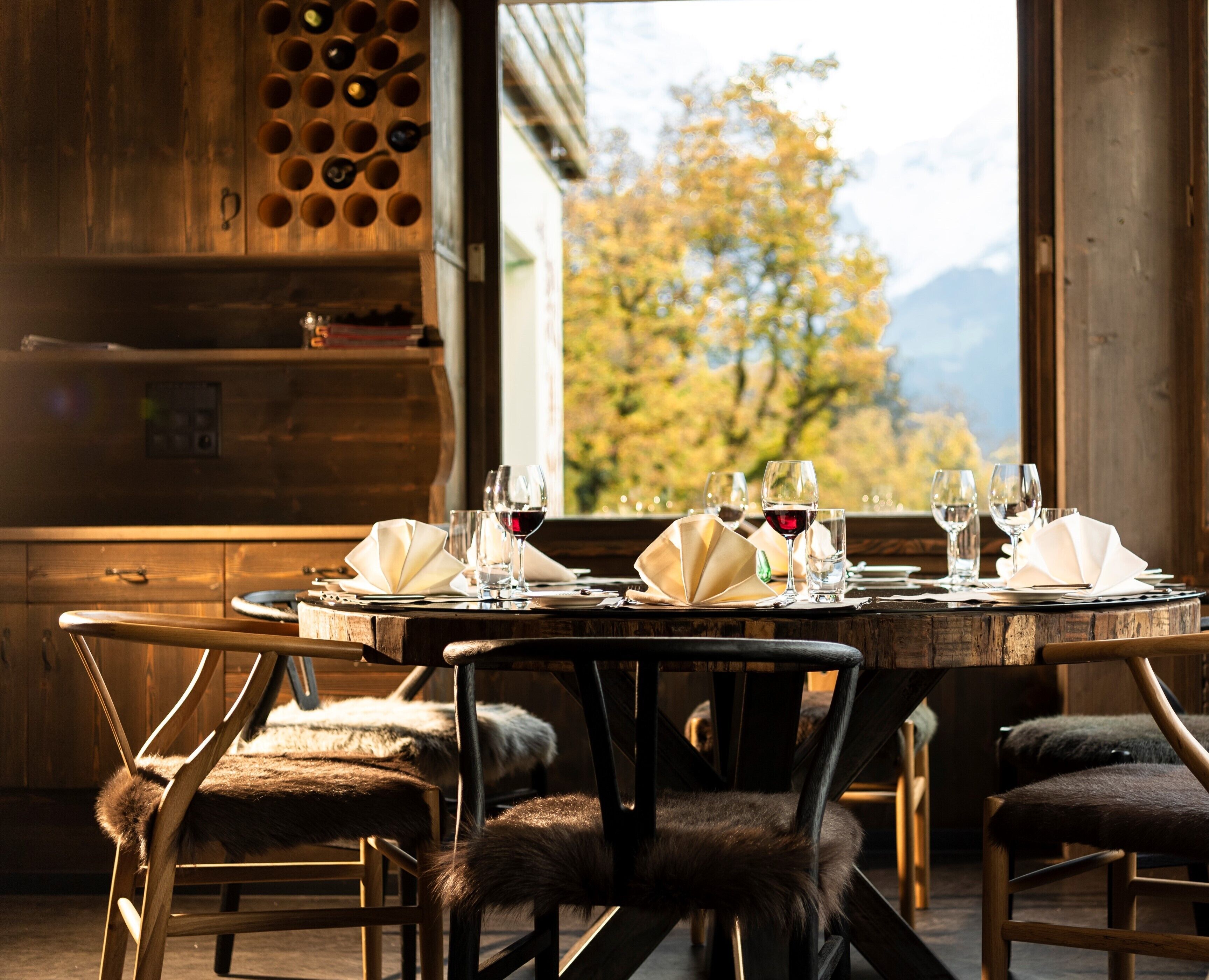 A dining table set for four with wine glasses and napkins, wooden chairs with fur throw seats, and a view of autumn trees through an open door.