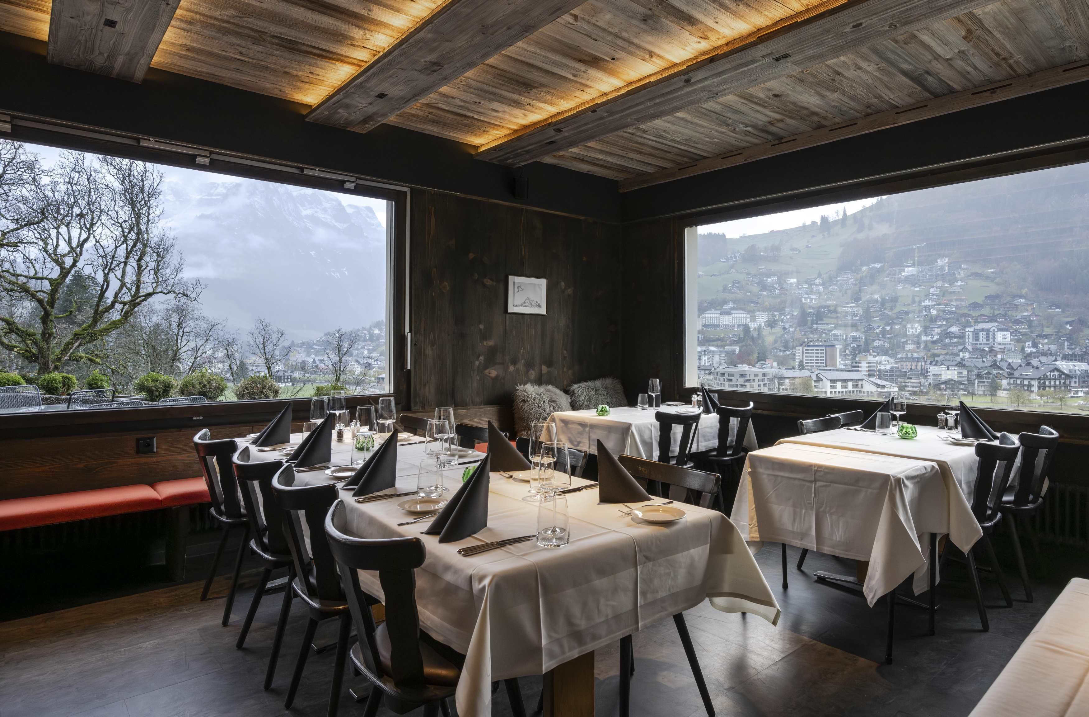 A restaurant with tables set for dining, large windows offering a city view, and a wooden ceiling.