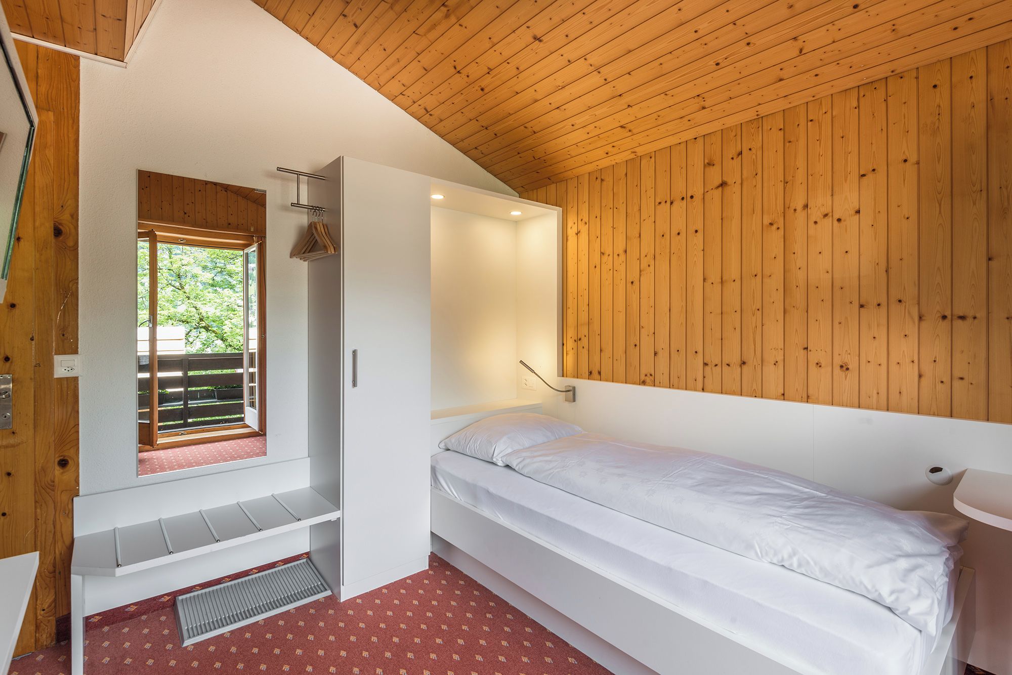 A bedroom with a wooden ceiling, a bed, a window with a view of greenery, and a built-in shelf.