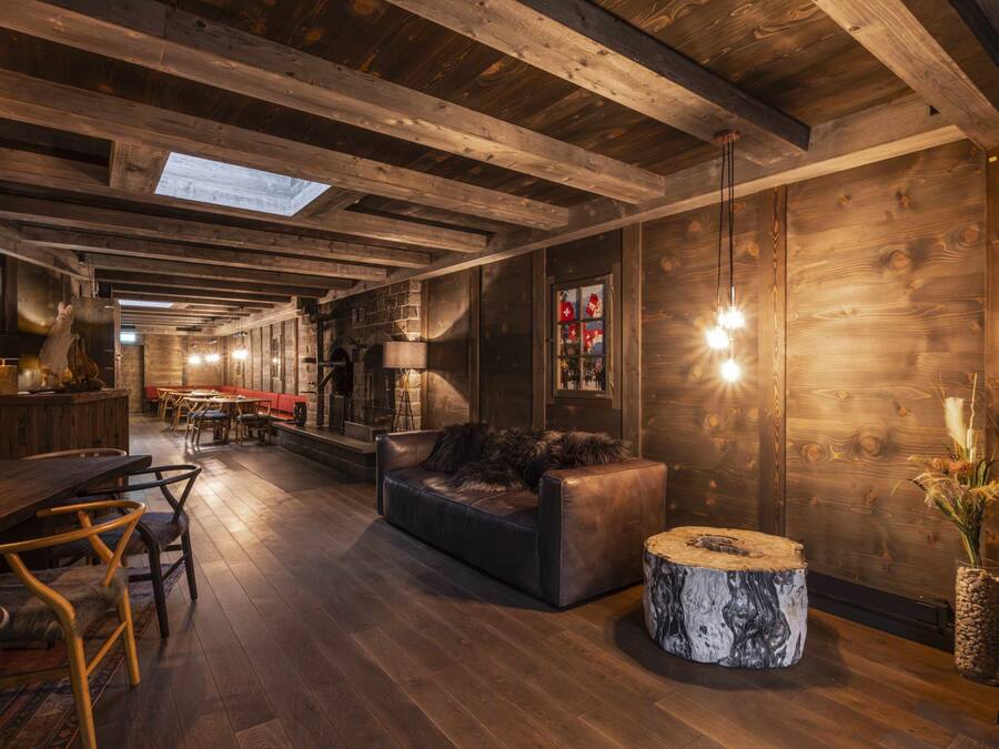 A rustic interior with wooden beams, a leather sofa, a round wooden table, and a bar area with stools.