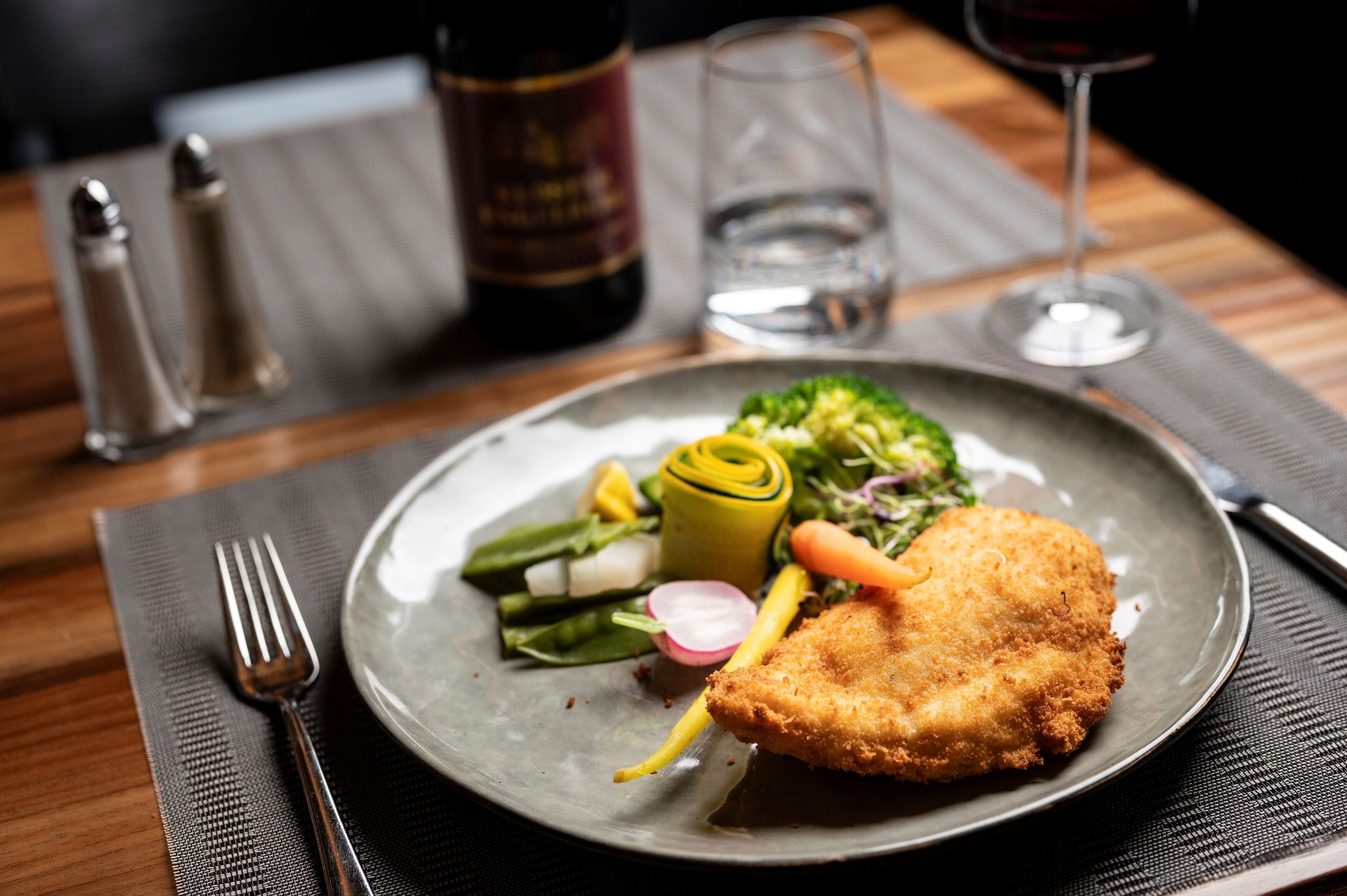 A plate with a breaded cutlet, vegetables, and a bottle of wine on a table.