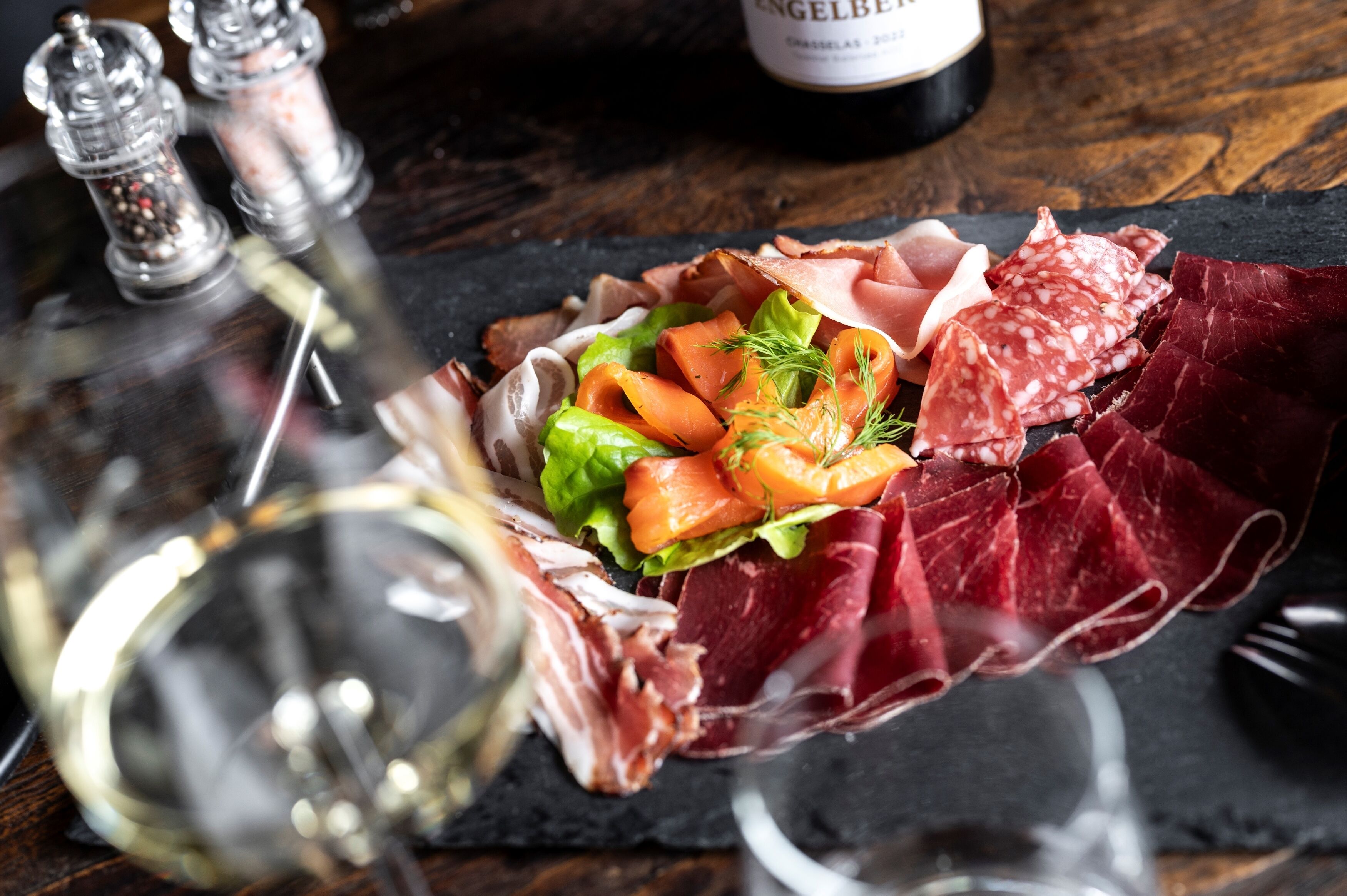 A charcuterie board with various meats, vegetables, and a bottle of wine on a dark slate table.