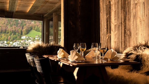 Una mesa de comedor dispuesta con servilletas blancas, copas de vino y vistas a un área boscosa a través de una ventana.