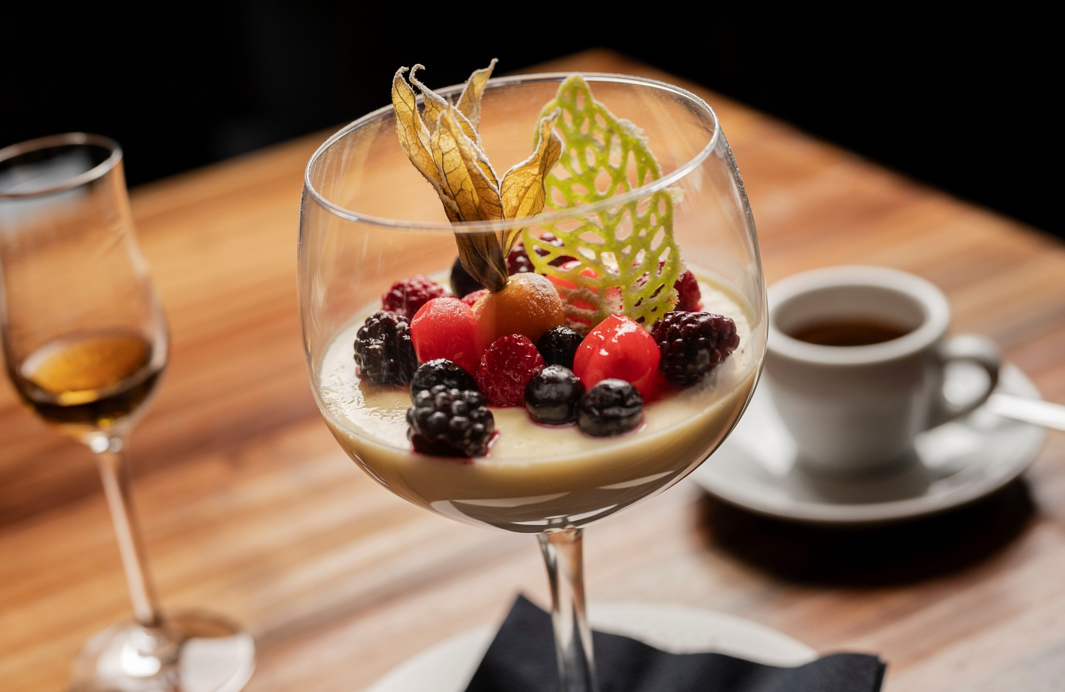 A dessert in a glass on a wooden table, accompanied by a cup of coffee and a glass of wine.