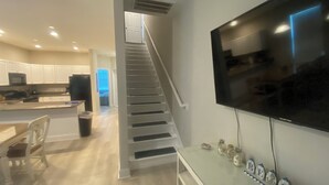 A modern kitchen with white cabinets, a dining area with a wooden table and chairs, a staircase leading to a second floor, and a mounted flat-screen TV on the wall. - RAISED BEACH HOUSE 1 BLOCK FROM BEACH (Myrtle Beach)
