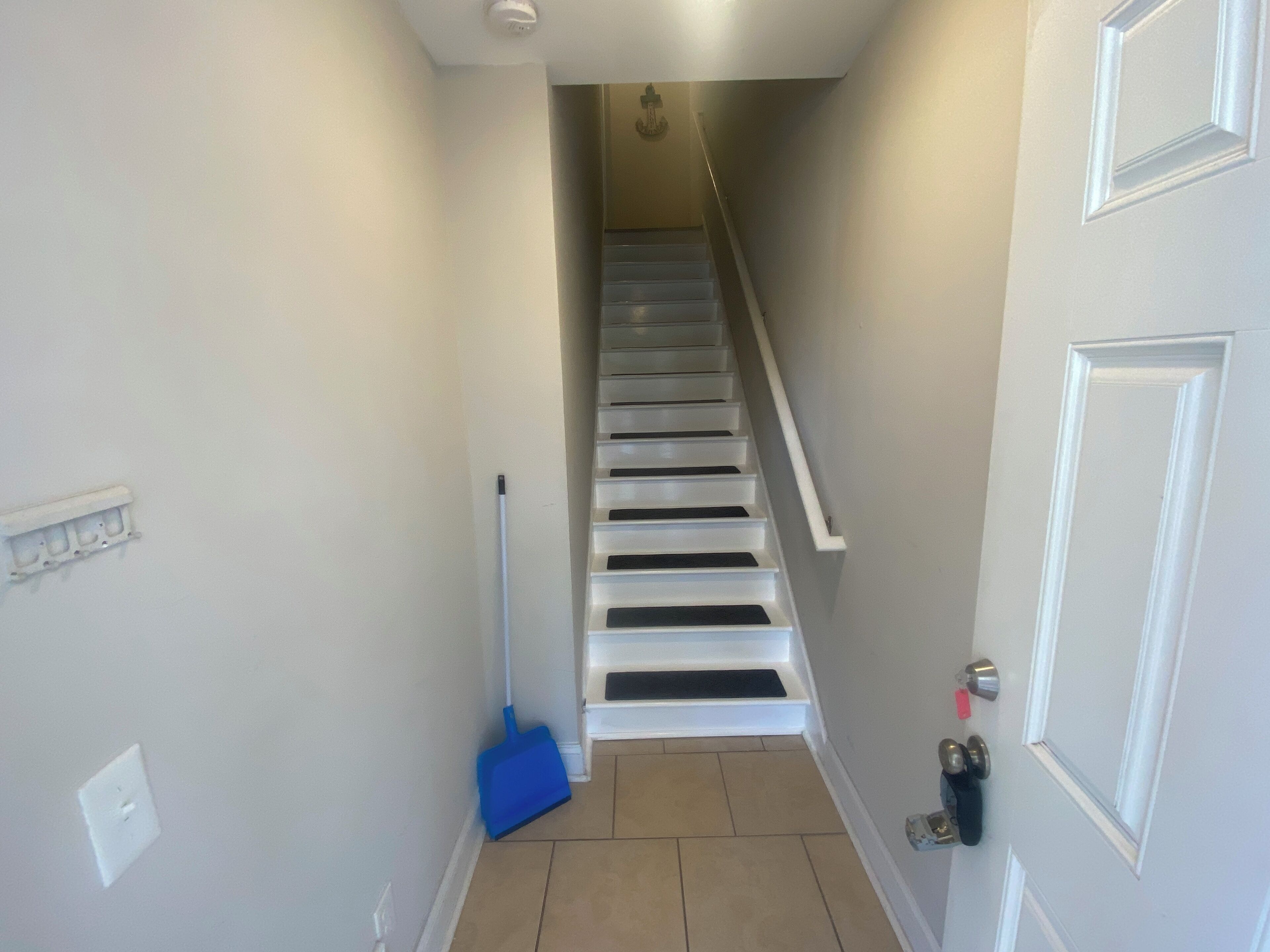 A narrow hallway with a white door on the right, a blue broom leaning against the wall, and a staircase leading to an upper floor.