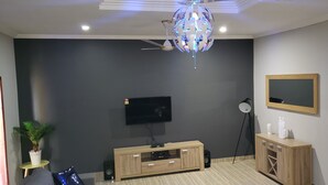 A modern living room with a flat-screen TV mounted on the wall, a wooden entertainment unit, a ceiling fan, and a decorative light fixture. - Candice serviced Apartment (Sunyani)