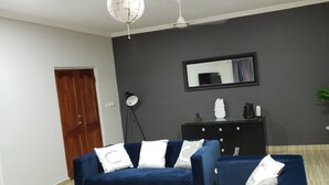 A living room with a dark blue sofa, a black entertainment unit, a mirror, a floor lamp, and a potted plant. - Candice serviced Apartment (Sunyani)