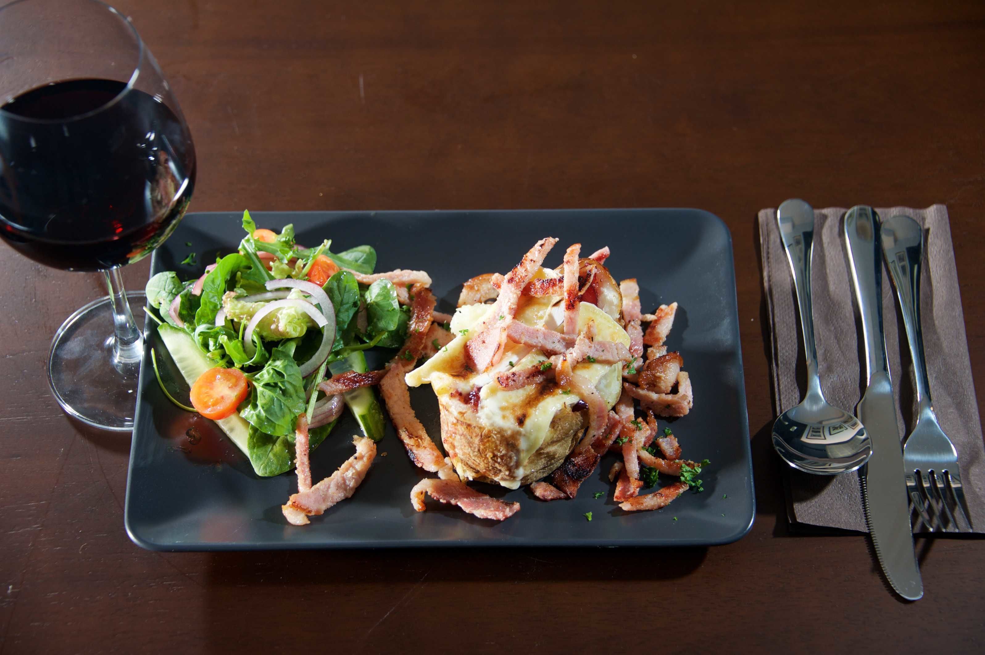 Un plato rectangular con una ensalada y un plato principal, acompañado de una copa de vino tinto y cubiertos sobre una servilleta.