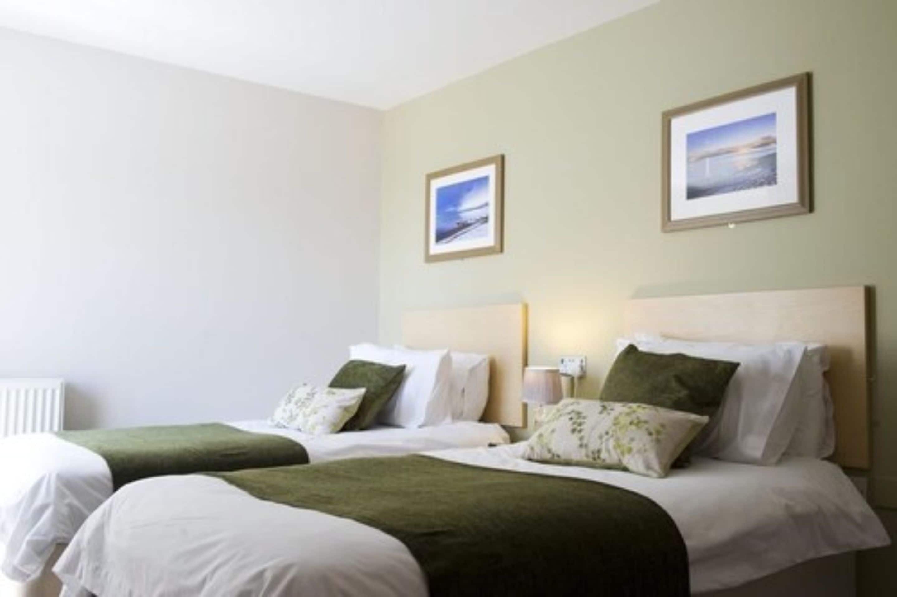 A hotel room with two beds, each with white linens and green throw blankets. There are framed pictures on the walls, a bedside lamp, and a radiator.