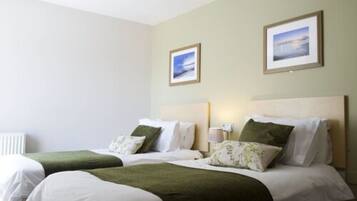 A hotel room with two beds, each with white linens and green throw blankets. There are framed pictures on the walls, a bedside lamp, and a radiator.