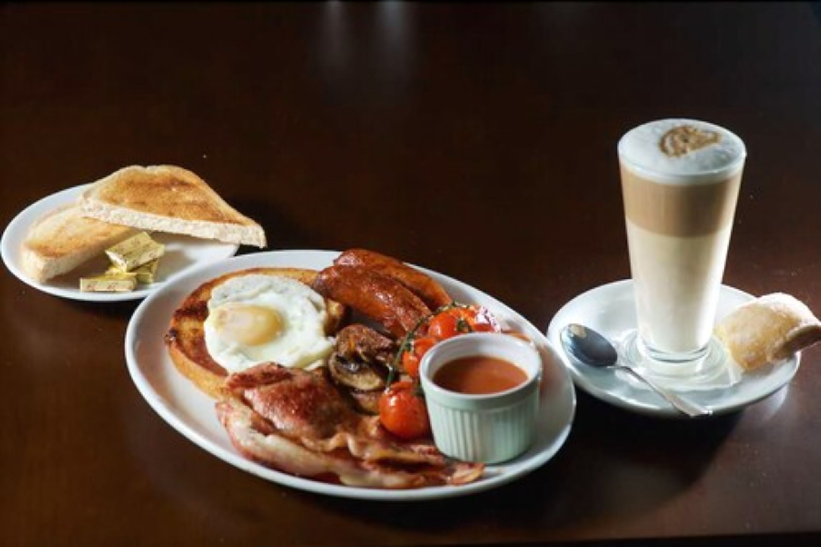 Une assiette de nourriture pour le petit-déjeuner comprenant des œufs, du bacon, des saucisses et des toasts, accompagnée d’une tasse de café et d’un petit bol de confiture.