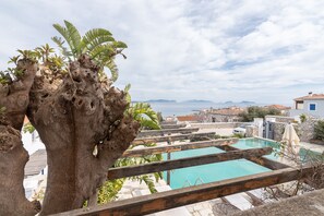 A pool area with a view of the sea and mountains, surrounded by stone buildings and tropical plants.