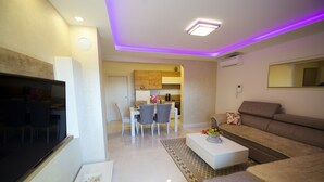 A modern living room with a flat-screen TV, a sofa, a coffee table, and a dining area with chairs and a kitchen in the background. - Apartments Leone (Baska Voda)