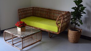 Un sofá de mimbre con un cojín verde, una mesa con superficie de vidrio y un jarrón de flores rojas, y una cesta tejida con una planta.