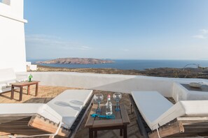 A terrace with white lounge chairs, a table with wine glasses and a bottle, and a view of the sea and distant hills.