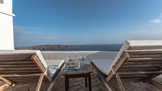 Eine Terrasse mit zwei Holzliegen, einem kleinen Tisch mit einer Flasche und zwei Gläsern, und Blick auf das Meer und das entfernte Land.