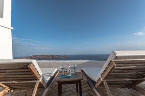 Une terrasse avec deux fauteuils de jardin en bois, une petite table sur laquelle se trouvent une bouteille et deux verres, et une vue sur la mer et le paysage lointain.