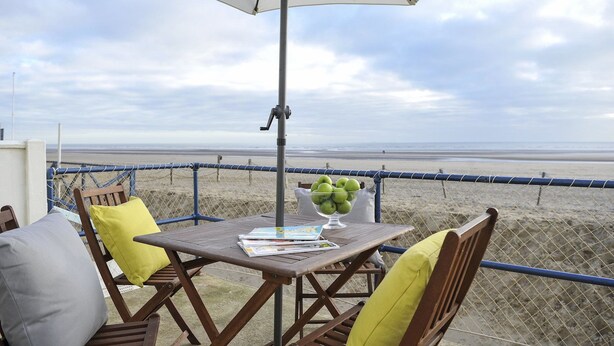 Une table avec un saladier de pommes vertes, un magazine et un parasol blanc sur un balcon donnant sur une plage.
