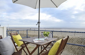 Une table avec un saladier de pommes vertes, un magazine et un parasol blanc sur un balcon donnant sur une plage.