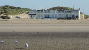 Un bâtiment situé sur la côte, doté d’un toit plat et de nombreuses fenêtres, et qui se trouve sur une plage de sable, avec des dunes en arrière-plan.