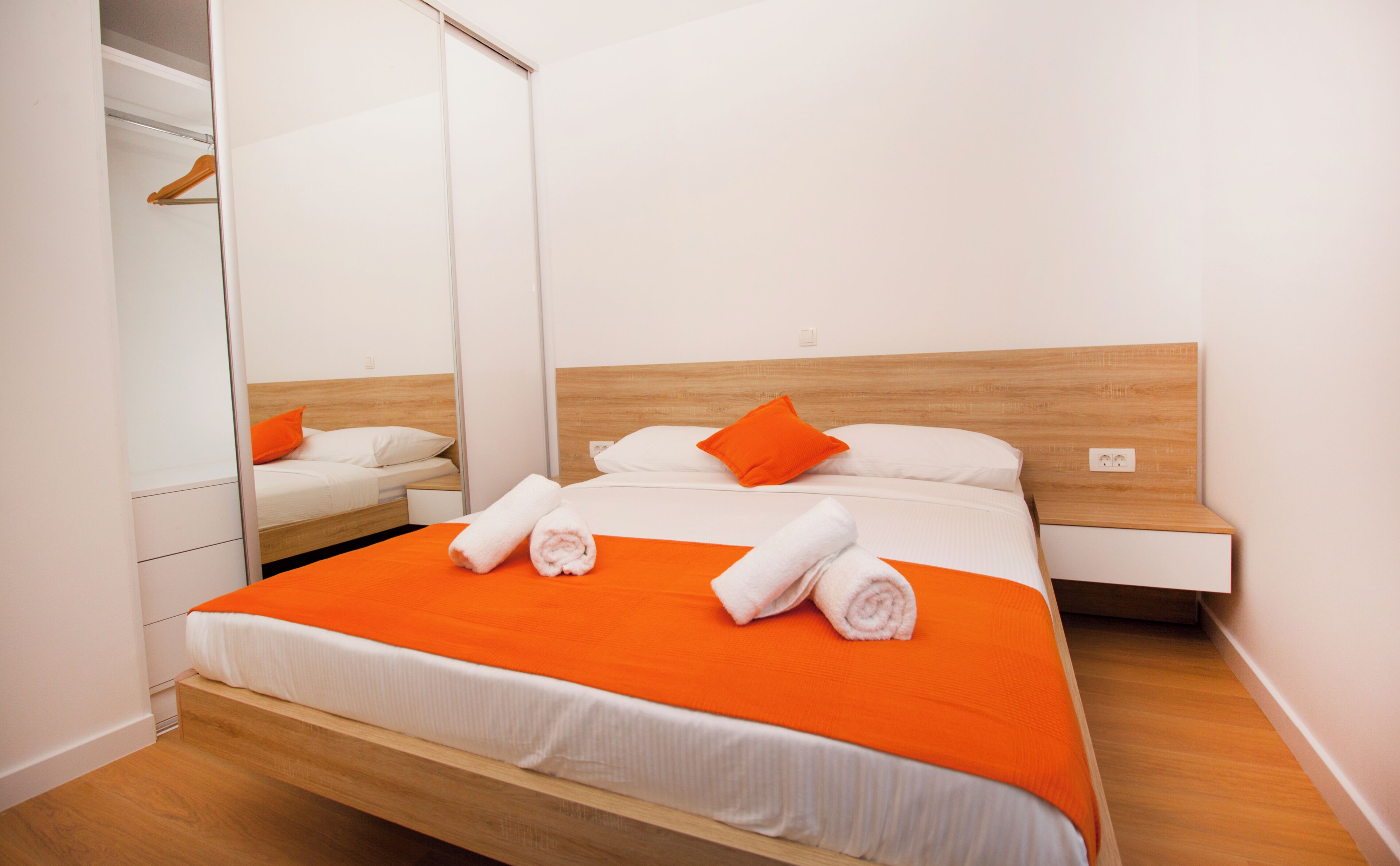 A modern hotel room with a wooden bed frame, white bedding, and orange accents.