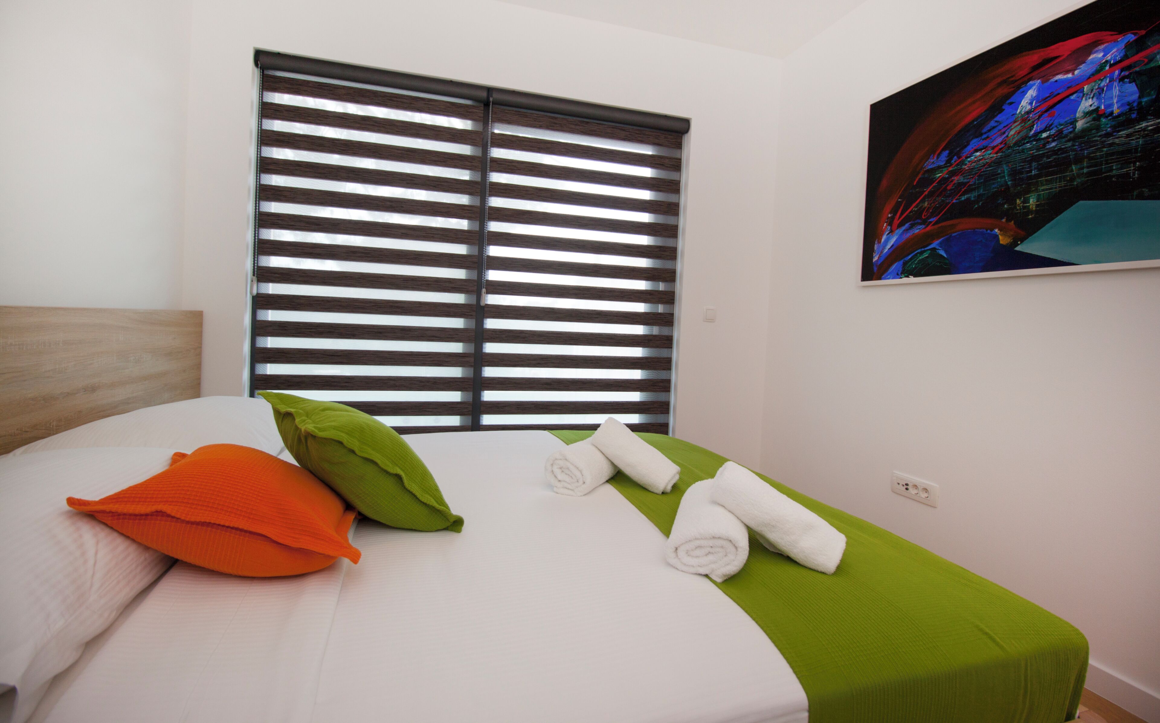 A hotel room with a bed, pillows, a window with blinds, and a painting on the wall.