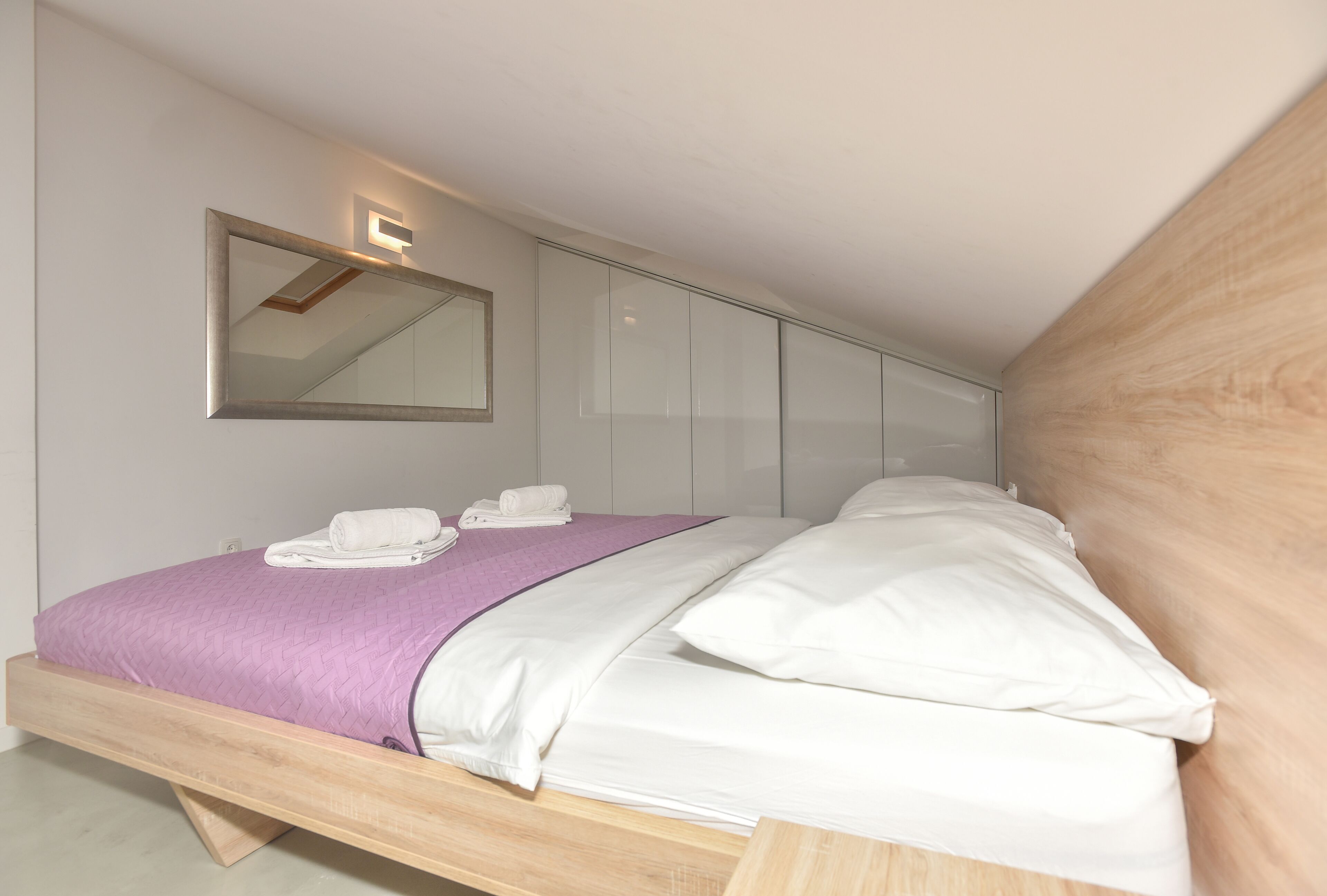 A modern bedroom with a wooden bed frame, a purple blanket, white pillows, and a wall-mounted mirror.
