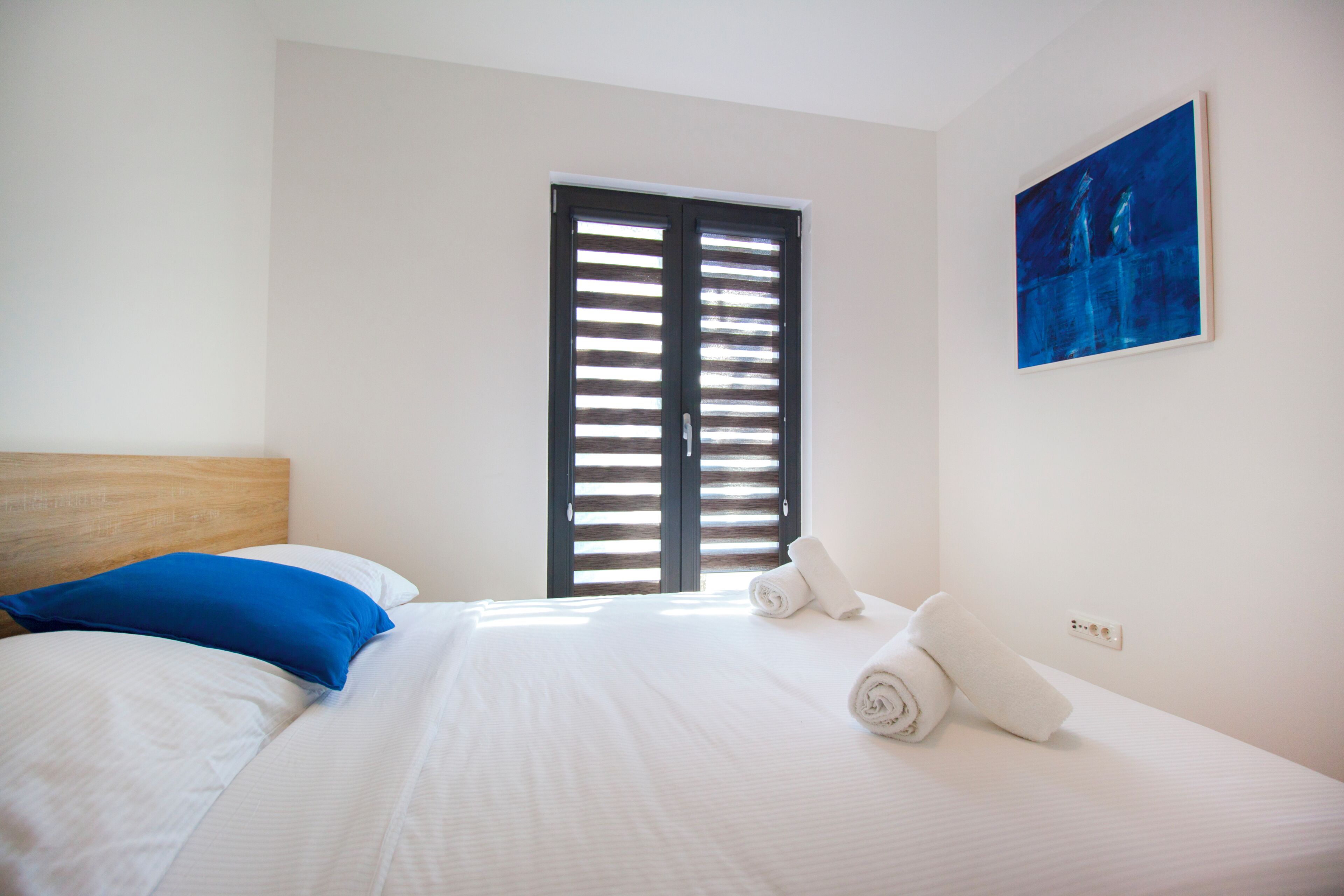 A hotel room with a bed, a blue pillow, a white pillow, a rolled white towel, a painting on the wall, and a window with blinds.