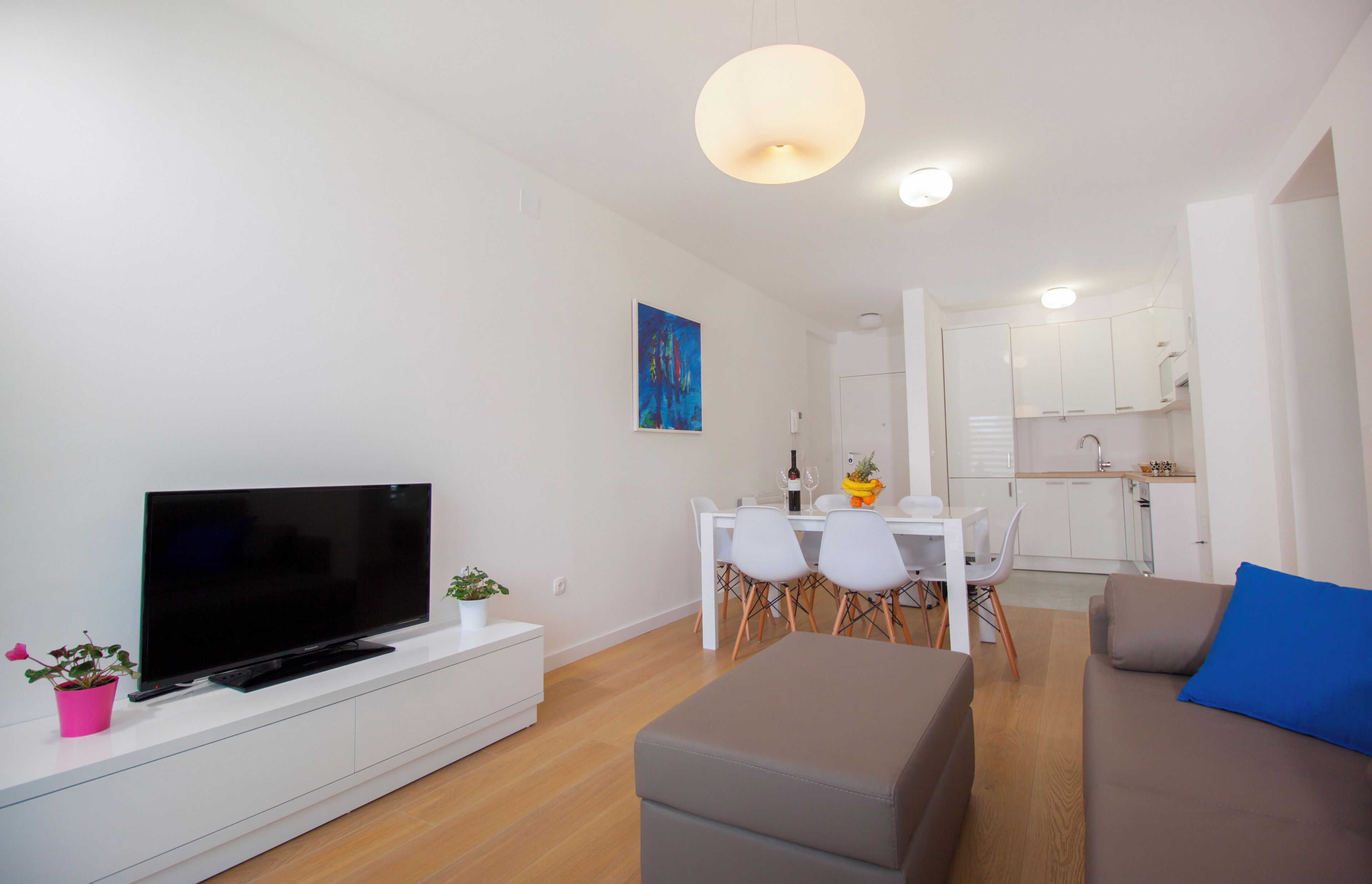 A modern living room with a flat-screen TV, a dining table with chairs, a kitchen area, and a sofa with a blue pillow.