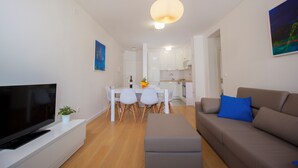 Ein modernes Wohnzimmer mit Flachbildfernseher, einer Couch, einem Esstisch mit Stühlen und einer Küche im Hintergrund.