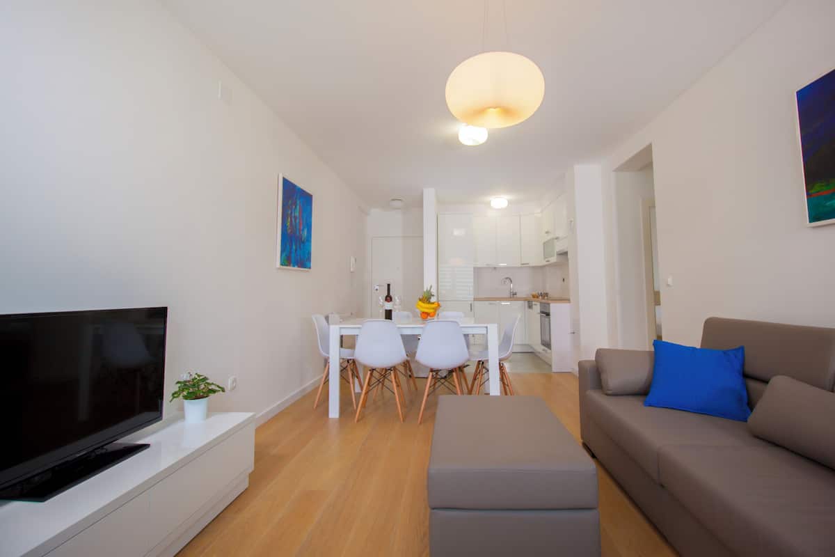 Een moderne woonkamer met een flatscreen televisie, een bank, een eettafel met stoelen en een keuken op de achtergrond.