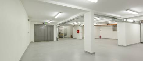 A spacious, modern indoor area with white walls and a polished concrete floor, featuring a central elevator and a fire extinguisher on the right.