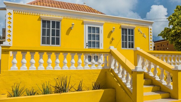 A yellow building with a red-tiled roof, white windows, and a white balustrade. - Old Aloe House (Savaneta)