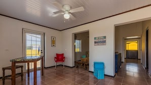 A spacious room with a ceiling fan, a wooden table, a red chair, and a blue storage bin. - Old Aloe House (Savaneta)