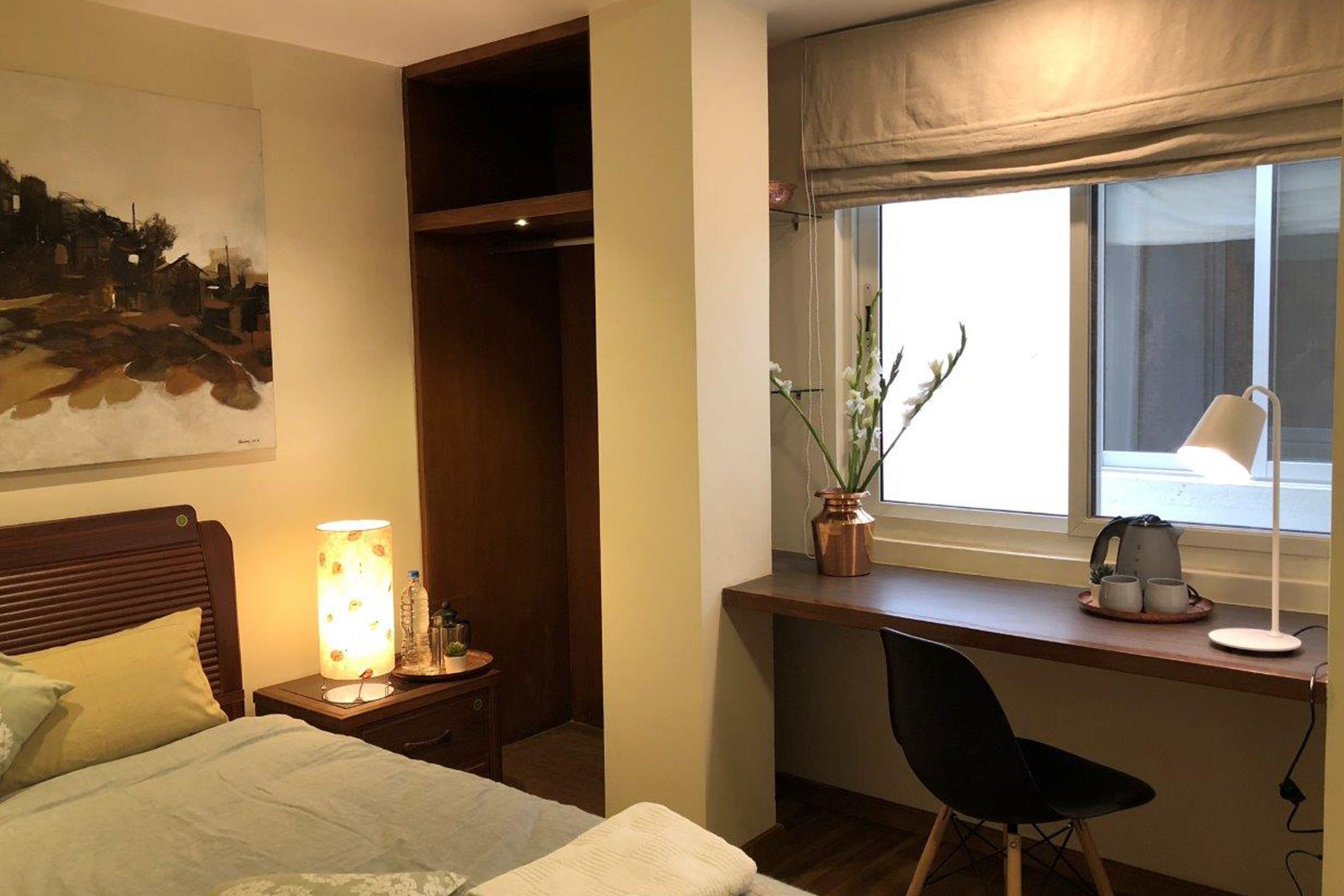 A bedroom with a bed, bedside table with a lamp, a desk with a chair, a window with curtains, and a potted plant.