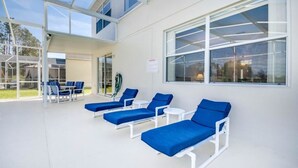A sunlit outdoor lounge area with white metal loungers and blue cushions, large windows, and a clear view of the surrounding greenery.