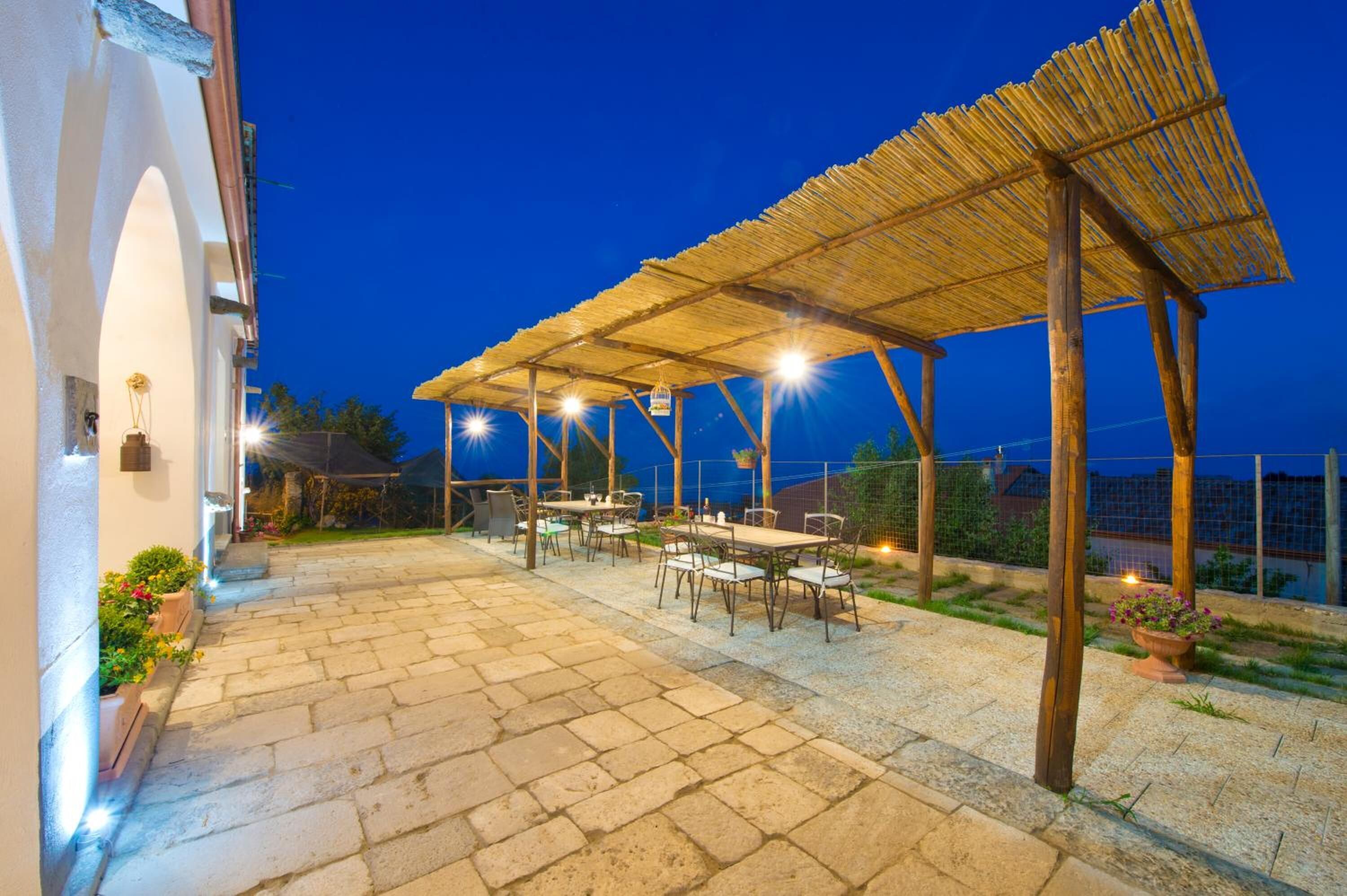 An outdoor patio area with stone flooring, wooden tables and chairs, and a thatched roof structure.