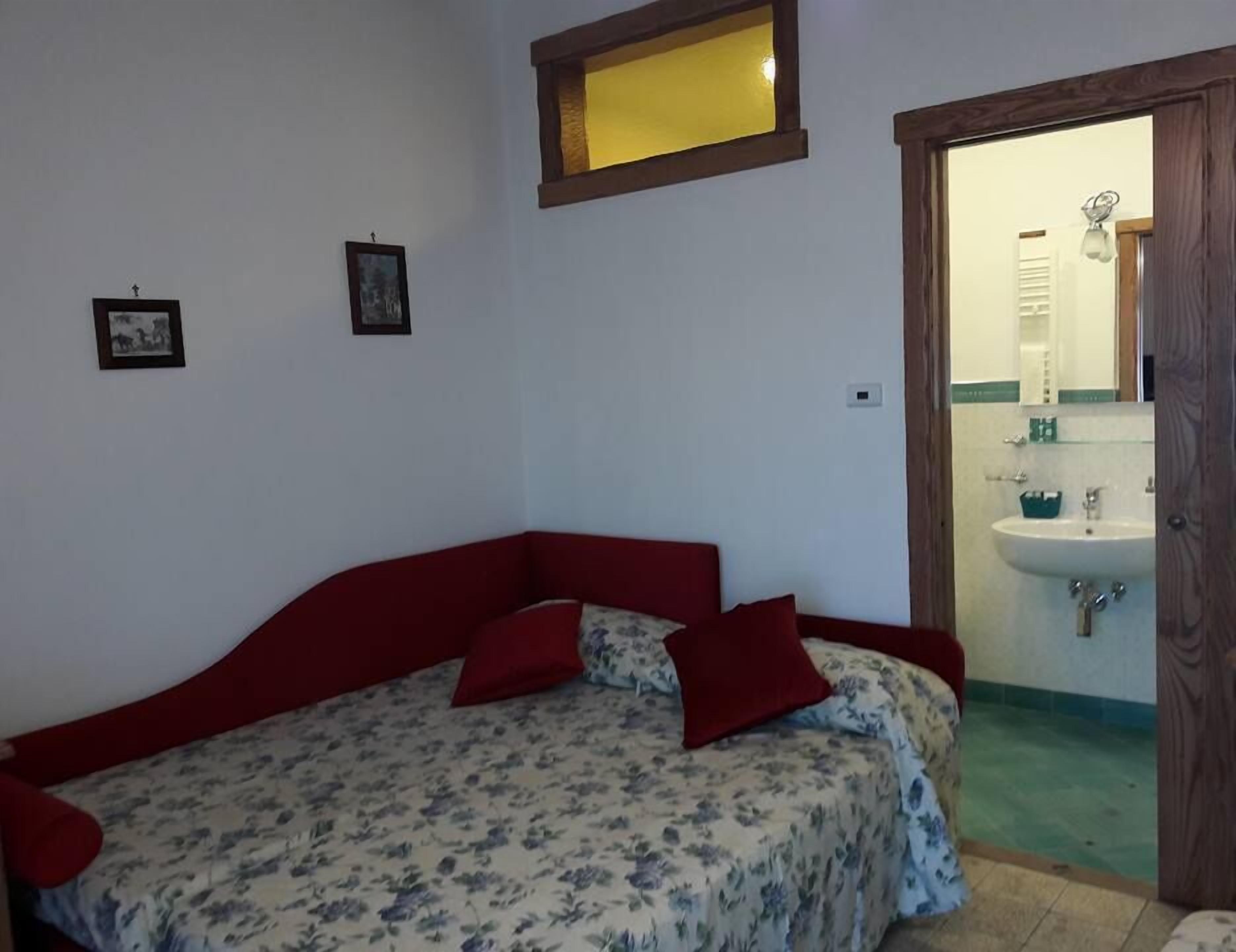 Un dormitorio con una cama roja, ropa de cama floral y una puerta de madera que da a un baño con lavamanos y espejo.