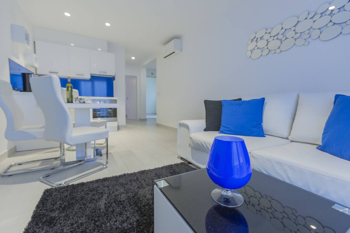 Un salon moderne avec un canapé blanc, des coussins bleus, une table basse en verre et un fauteuil blanc.