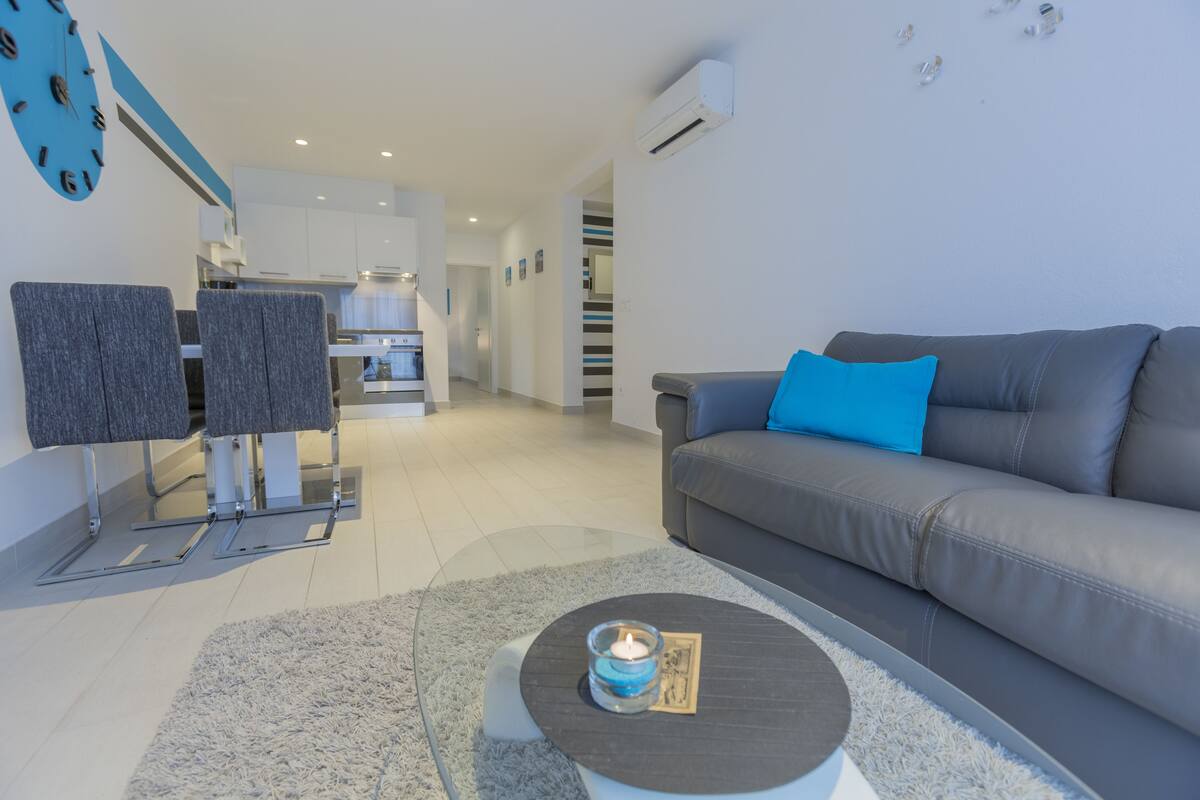 Un salon moderne avec un canapé gris, un coussin bleu et une table basse en verre sur laquelle est posée une bougie.