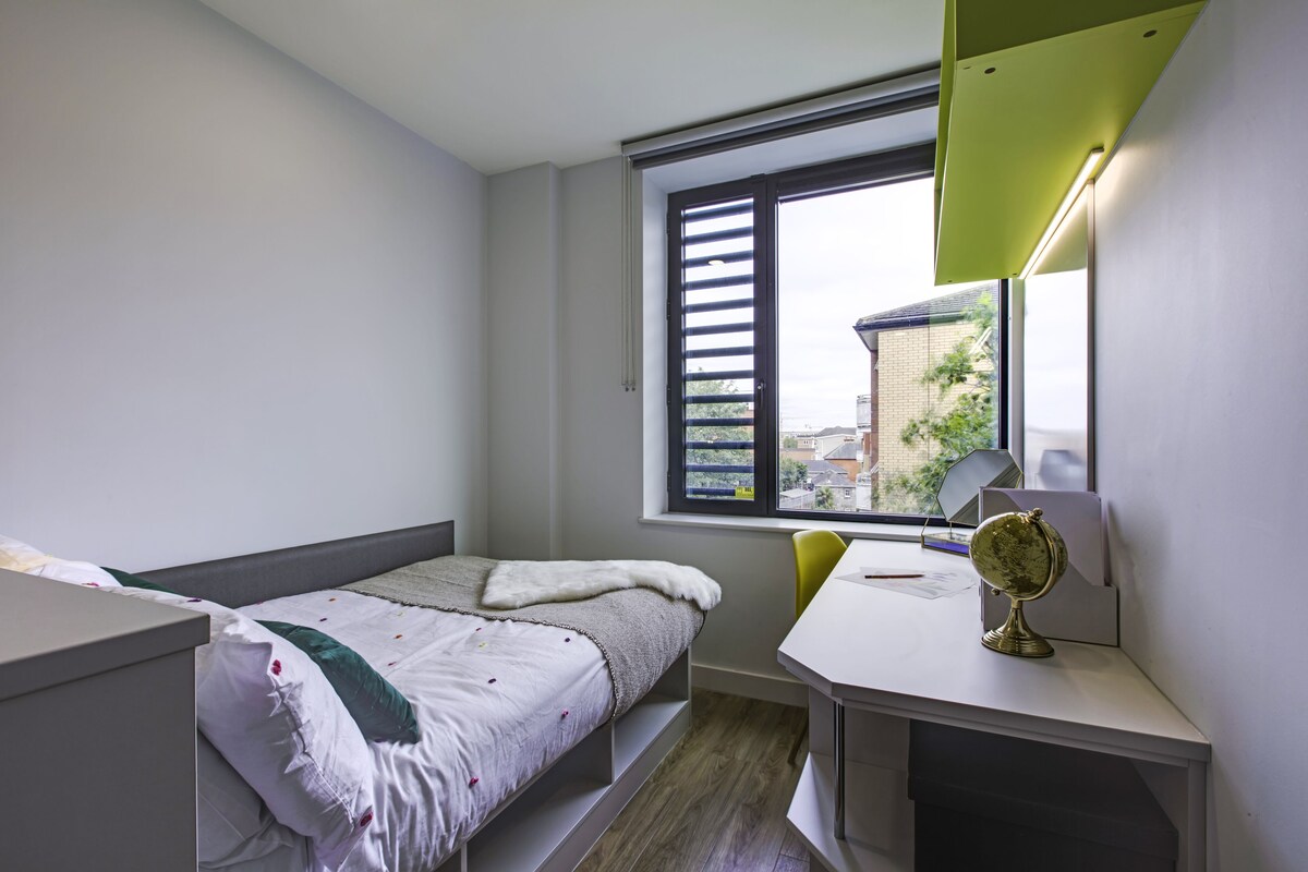 A bedroom with a bed, a desk, a chair, a window with blinds, and a mirror.